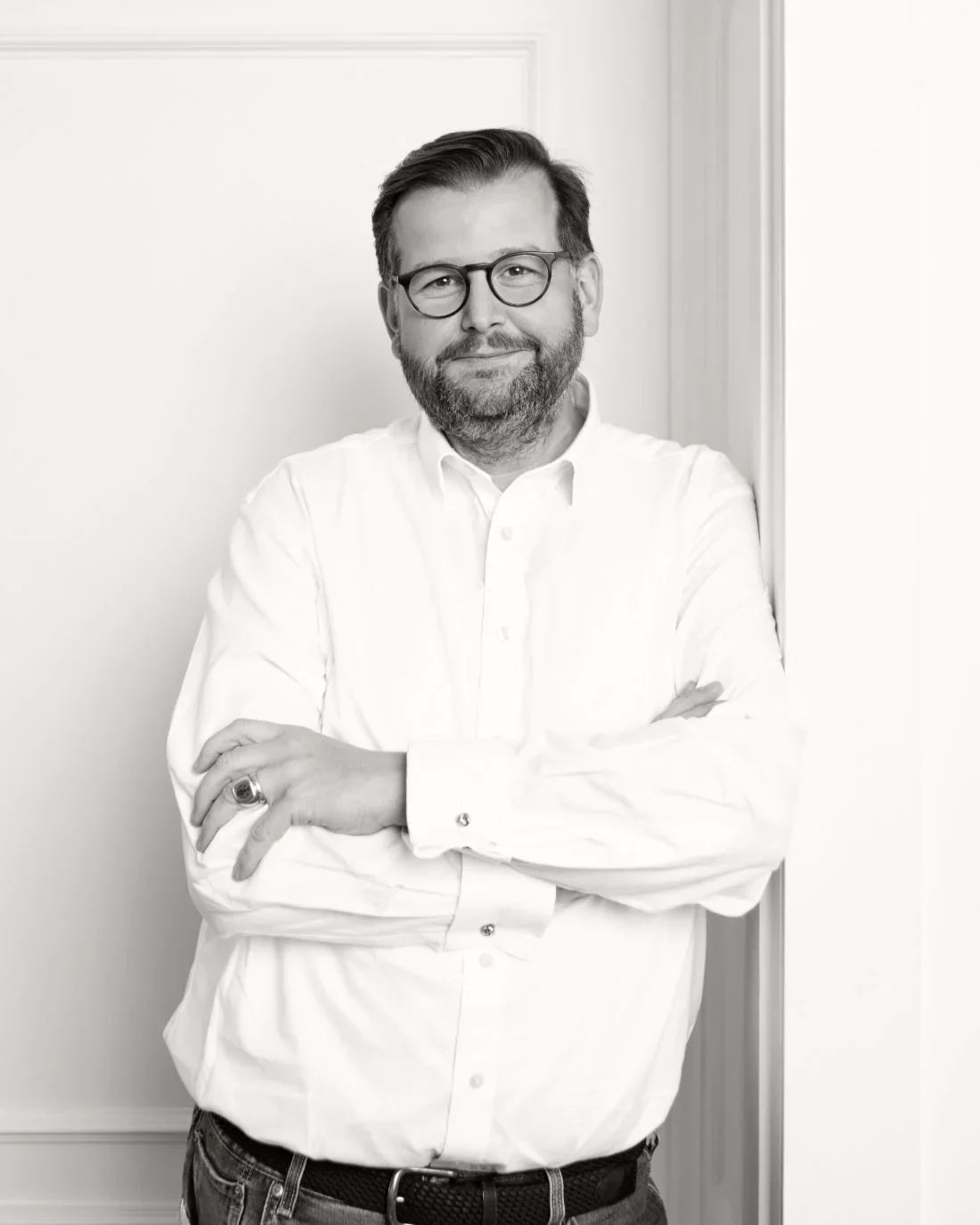 Schwarz-Weiß-Foto eines Mannes mit Brille, Bart und gepflegtem Haar, der lächelnd mit verschränkten Armen vor einer weißen Wand steht.